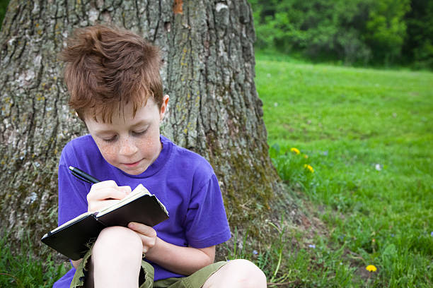 Beautiful little boy sitting in grass outside, writing in a notebook or journal.