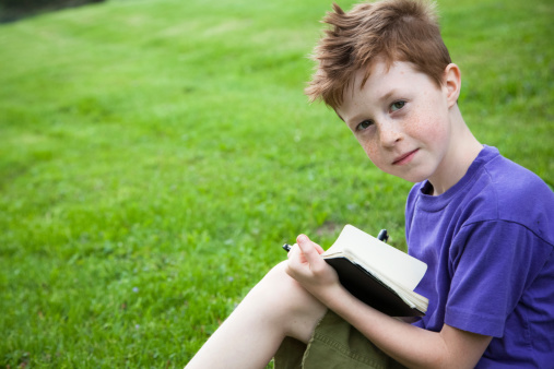 Beautiful little boy sitting in grass outside, writing in a notebook or journal.