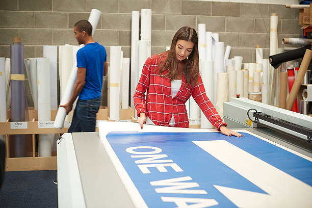 young woman working at a digital printing company A young woman is working on a digital printing machine . she is printing out a large one way sign . A colleague is choosing more vinyl for the next job. digital printing house stock pictures, royalty-free photos & images