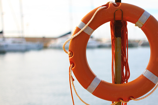 A bright orange life preserver (life ring) in the foreground with a pale morning sea, sky and sailboats in the blurred background. Copy space in the pale water.