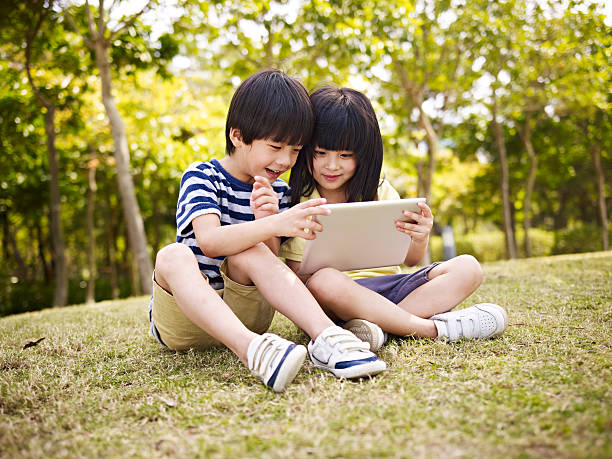 two asian children using tablet outdoors little asian girl and boy sitting on grass using digital tablet outdoors in a park. korean boy stock pictures, royalty-free photos & images
