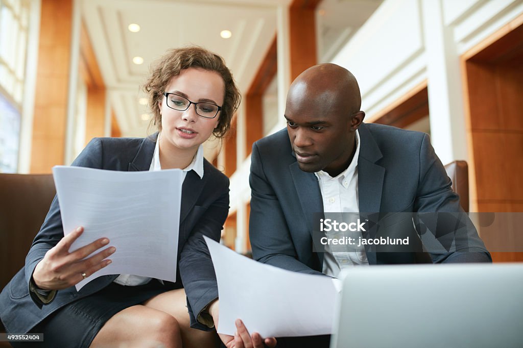 Two business partners sitting in cafe and discussing contract. Portrait of young business man and woman sitting in cafe and discussing contract. Diverse businesspeople meeting in hotel lobby reading documents. Examining Stock Photo