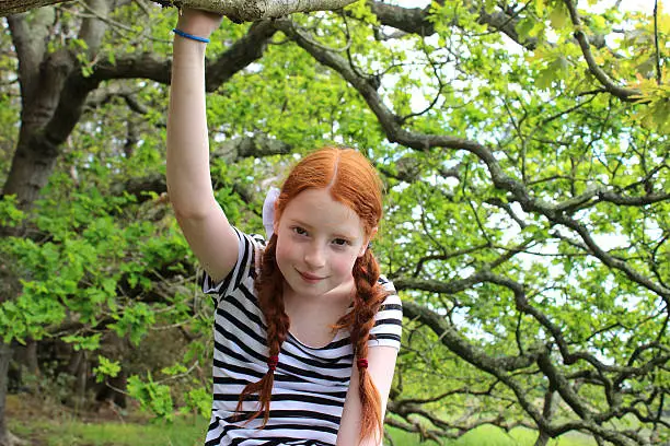 Photo showing a young tomboy girl sitting on the branch of a large English oak tree that she has just climbed up, in a woodland setting.