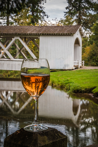 Wine glass featuring Rosé, pink hued Oregon Wine with inverted image of the Covered Bridge in Stayton. Also known as the Thomas Creek Bridge. Spanning Thomas Creek, seen while on a driving tour of Linn County Covered Bridges. Serene setting in a park.  Still waters in Creek reflecting the bridge and trees. Vertical Close up shot highlighting reflections in water and wine glass. Artistic and Illustrative. No people, nobody.