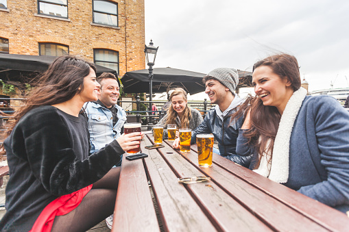 Group of friends enjoying a beer at pub in London, toasting and laughing. They are seated outside at a wood table, wearing winter clothes. Friendship and lifestyle concepts.