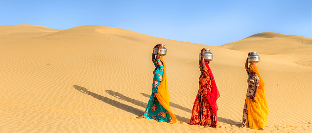 Indian women crossing sand dunes and carrying on their heads water from local well, Thar Desert, Rajasthan, India. Rajasthani women and children often walk long distances through the desert to bring back jugs of water that they carry on their heads. Panoramic view.