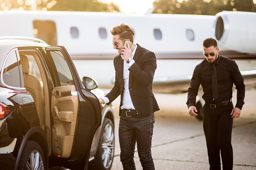 Two well dressed men arriving at the airport track to catch a flight. Man on the left is closing the car door while talking over mobile phone and his friend is next to him. Private jet airplane is in the background.