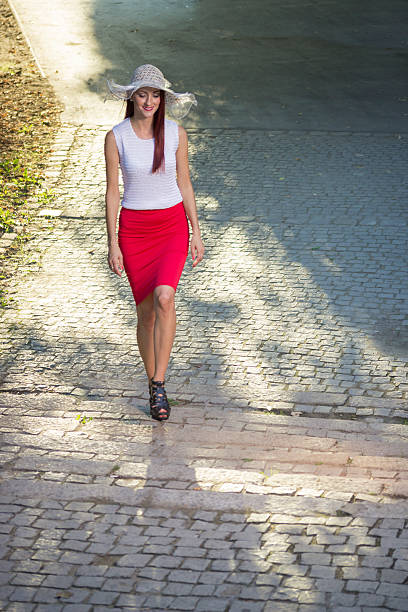 Young elegant girl in 20s walking in park. Red dress, whiter shirt and hat.