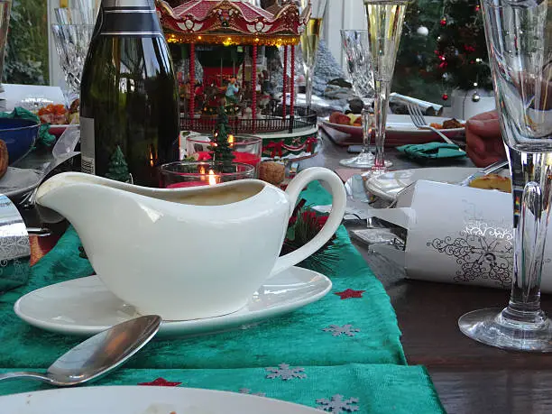 Photo showing a white porcelain gravy boat on a Christmas dinner table, where it is accompanied by seasonal decorations, wine glasses, a bottle of champagne / sparkling wine, dinner plates and a toy carousel.  A green velour table runner adds colour to the arrangement.