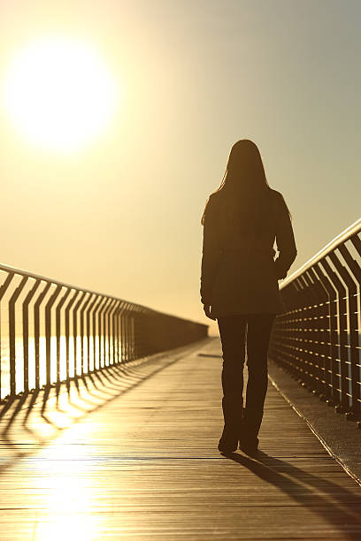 Sad woman silhouette walking alone at sunset Sad woman silhouette walking alone on a bridge on the beach in winter at sunset walking beach women sadness stock pictures, royalty-free photos & images