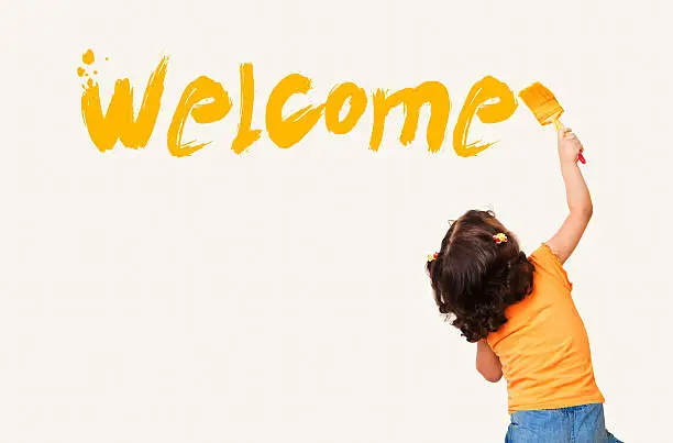 Cute little girl writing "Welcome" with painting brush on wall background