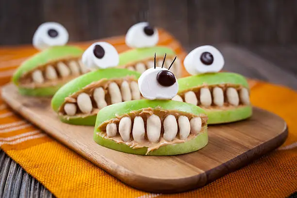 Spooky halloween edible apple monsters healthy natural dessert. Horror party decoration delicious snack. Homemade cute cyclop mouth with teeth and peanut butter on dark vintage wooden table background.