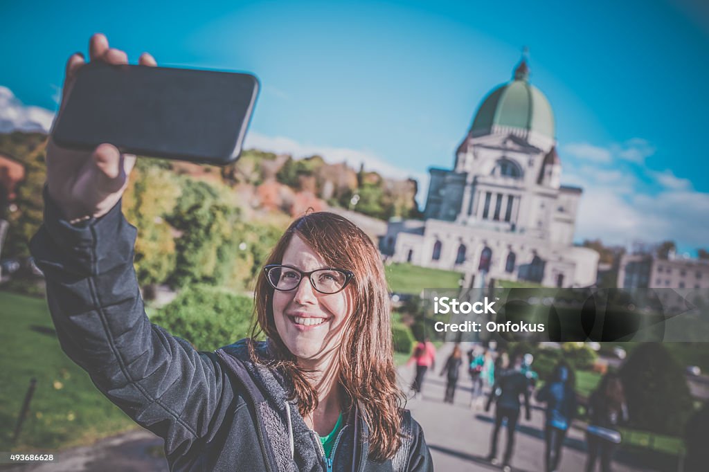 Woman Doing Selfie in Front of Saint Joseph's Oratory, Montreal DSLR picture of a woman tourist doing a selfie in front of the St Joseph Oratory in Autumn, Montreal, Quebec. It is a beautiful fall day. The man is happy and smiling. The Saint Joseph's Oratory is a Roman Catholic minor basilica and national shrine on Westmount Summit in Montreal, Quebec. It is Canada's largest church. A retro filter was applied to the picture. 2015 Stock Photo