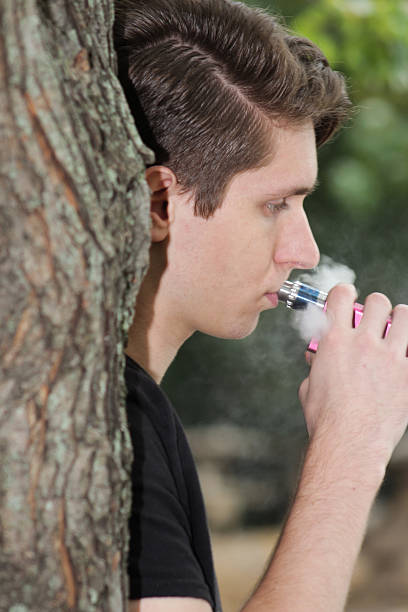 istock Adult Male Leans on Tree and Puffs Electronic Vaping Device 493720320