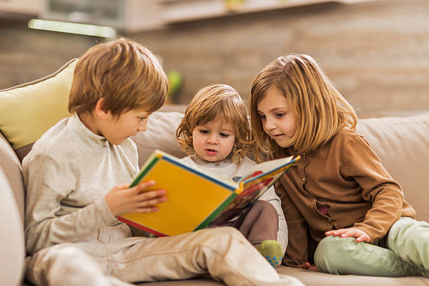 Children reading picture book at home. Little boy storytelling to his brother and sister at home. boy and girl reading stock pictures, royalty-free photos & images