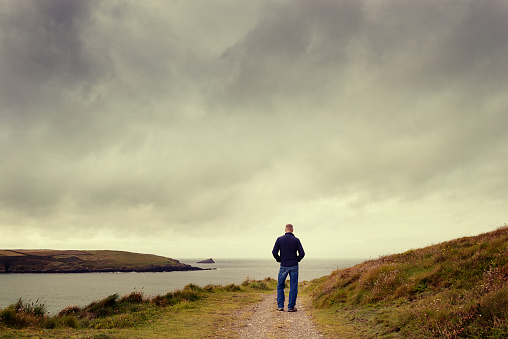 Man stood on a coastal footpath, alone, looking out over an overcast sky.