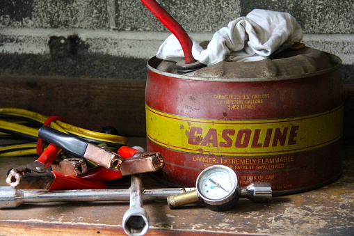 This is an old antique gasoline can with a rag, jumper cables, tire gauge and lug wrench set on a wooden shelf.