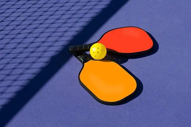 Photo of Pickleball - 2 paddles and ball sitting near net shadow