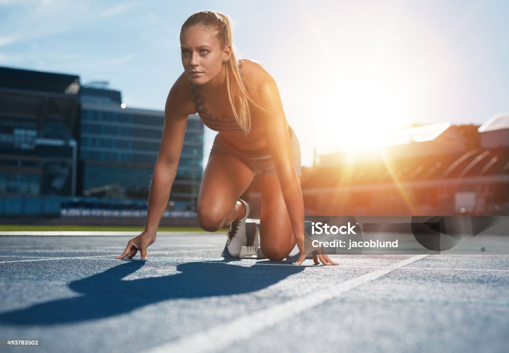 Running with determination Fit and confident woman in starting position ready for running. Female athlete about to start a sprint looking away. Bright sunlight from behind. Sprinting Stock Photo