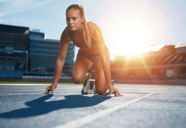 Running with determination Fit and confident woman in starting position ready for running. Female athlete about to start a sprint looking away. Bright sunlight from behind. runner on start pose stock pictures, royalty-free photos & images