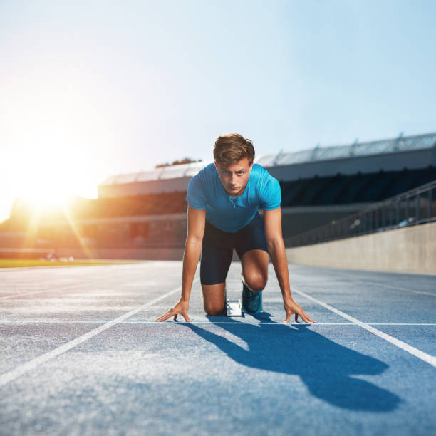 Fit and confident sprinter at starting blocks Fit and confident man in starting position ready for running. Male athlete about to start a sprint looking at camera with bright sunlight. mens track stock pictures, royalty-free photos & images