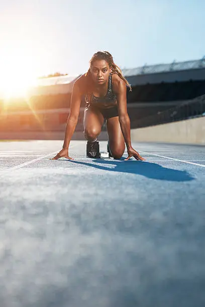 Vertical shot of young female sprinter taking ready to start position facing the camera.  Woman athlete in starting blocks with sun flare.