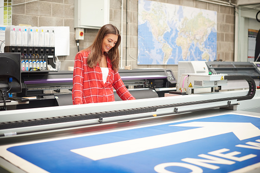 A young woman is working on a digital printing machine . she is printing out a large one way sign . ** background map has been modified**