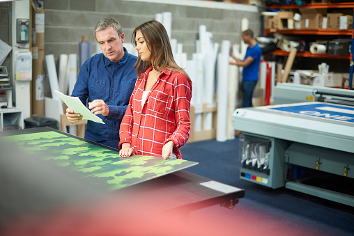 A young woman is working on a digital printing machine . her boss is explaining the machine or the job that she is doing . In the background rolls of vinyl can be seen , and a young co-worker is cutting out some vinyl lettering   