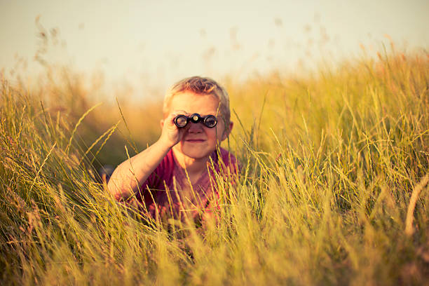 Young Boy Looking Through Binoculars Hiding in Grass A young boy is looking through binoculars while hiding in the tall grass of an English field. He could be spying on or searching for something newsworthy. Image taken in the sunset light and with shallow depth of field. hiding place stock pictures, royalty-free photos & images