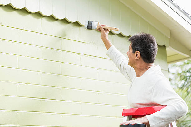 Mature Hispanic man (50s) standing on ladder, painting house.