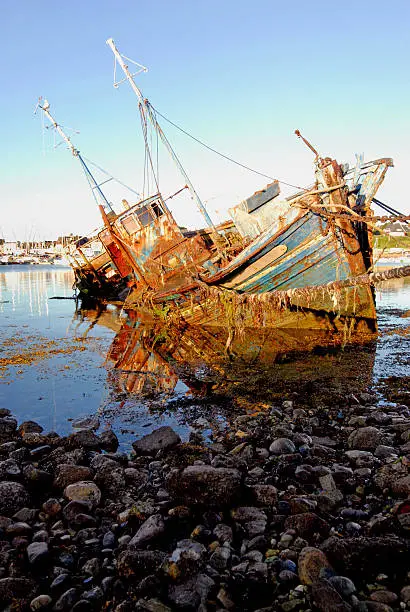 A rotting hulk of an old French fishing trawler lies on its side in a French west coast port whilst weed and barnacles accumulate on its hull and old mooring lines