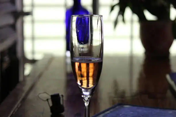 This image shows a glass of rose champagne against a well-lit dark counter/bar, and blue glass container to add another dimension of color. There are two other images of this setting. The other images show a vertical take on the setting, showing the entire glass with stem and more of the counter (called Glass of Rose Champagne / Sparlking Rose 1 of 3, Glass of Rose Champagne / Sparlking Rose 3 of 3).