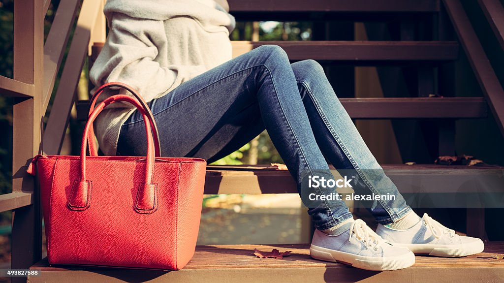 Girl on the stairs with a big red fashionable handbags Girl sitting on the stairs with a big red super fashionable handbags in a sweater jeans and sneakers on a warm summer evening. warm colors Women Stock Photo