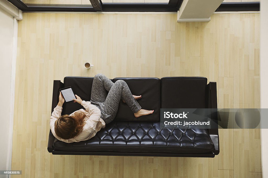 Young lady relaxing on a couch using digital tablet Top view of young lady sitting on a couch using digital tablet. Female relaxing on sofa with a e-reader in her apartment. Flooring Stock Photo