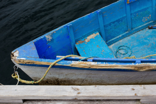 A weathered, blue, wooden dinghy (skiff, punt) rowboat tied with yellow rope to a floating dock slip on the coast of Maine, with gentle water waves.