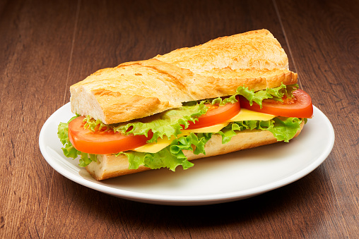 Tomato, cheese and salad sandwich from fresh baguette on white ceramic plate on dark wooden table background