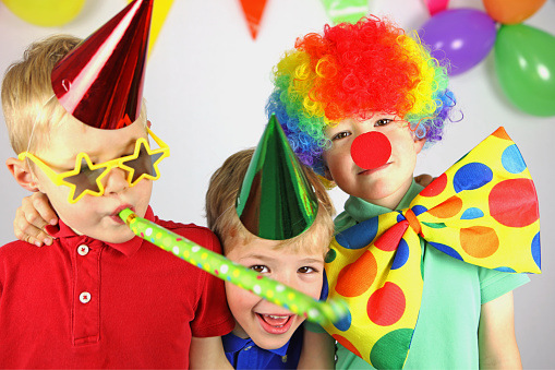 Three boys in colorful party clothing are celebrating a birthday. One child is wearing a colorful clown wig, a red nose, and a jumbo bow tie, his twin brother is wearing a red party hat and funny star-shaped sunglasses and is blowing a party blower. The older boy with a green party hat and a blue t-shirt is smiling and looking towards the camera. Colorful party flags and balloons can be seen behind the children.