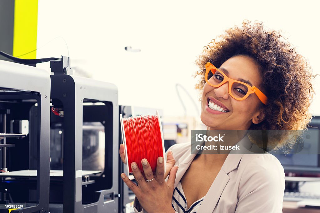 3D printer office Mixed race young woman wearing 3D printed glasses, sitting in a 3D printer office, holding red filament in hands and smiling at the camera. 3D Printing Stock Photo