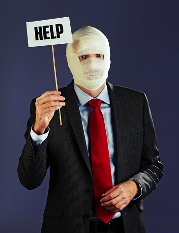 A suited businessman has his head and face obscured by mummy-like bandages, preventing him speaking. He holds up a sign saying simply 