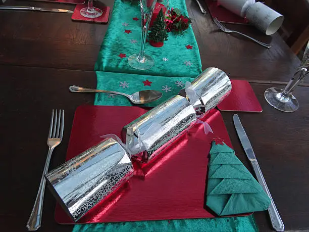 Photo showing a festive place setting on a Christmas dinner table, with some shiny silver crackers, a green table runner, a red placemat and a tree paper napkin, topped with a star.