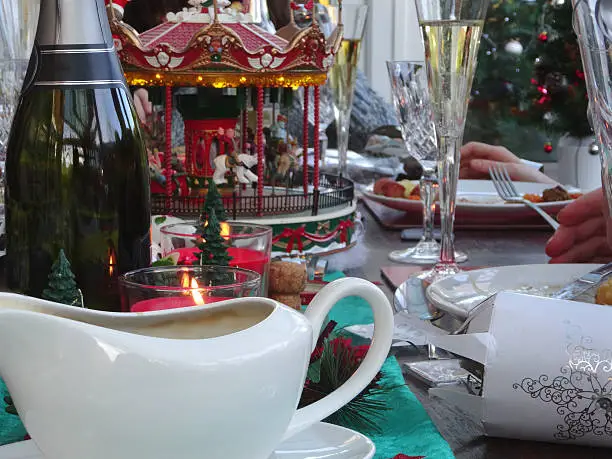 Photo showing an attractive Christmas dinner table and its decorations, which include a toy carousel roundabout ride, and lots of silver crackers.  Also on the table is a white china gravy boat and a bottle of sparkling wine.