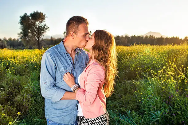 Attractive young couple enjoying a passionate kiss in the evening light against a backdrop of a colorful yellow field of rapeseed flowers.