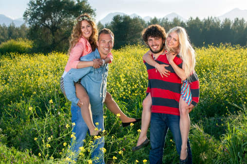 Happy young couples enjoying nature as the two smiling young men give their attractive girlfriends a piggy back ride through a yellow rapeseed field.