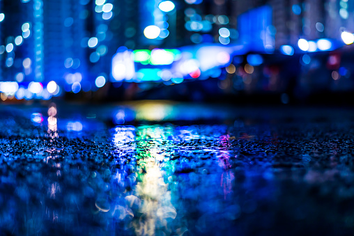 Night cityscape, colored lights reflected in the wet asphalt after rain. View from the level of asphalt, in blue tones