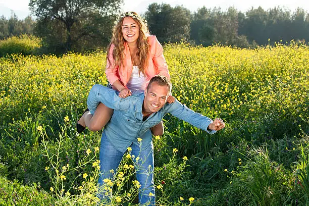 Playful loving young couple riding piggy back as they frolic around in a colorful yellow field of rapeseed on a sunny summer day.