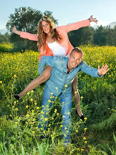 Happy lively young couple riding piggy back waving their arms and laughing as they enjoy a relaxing day in farmland in a yellow rapeseed field.