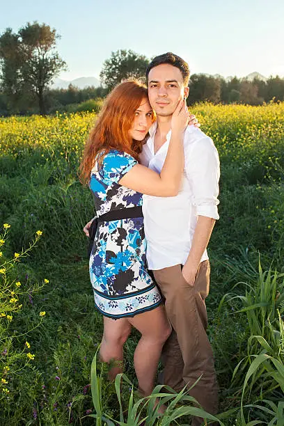 Affectionate young couple in the countryside standing in a field of yellow rapeseed with the girls arms around her boyfriends neck as though interrupted in a kiss  both looking at the camera.