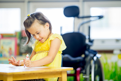 A happy little girl with cerebral palsy is sitting at a desk at an elementary school and is coloring a picture with a marker.