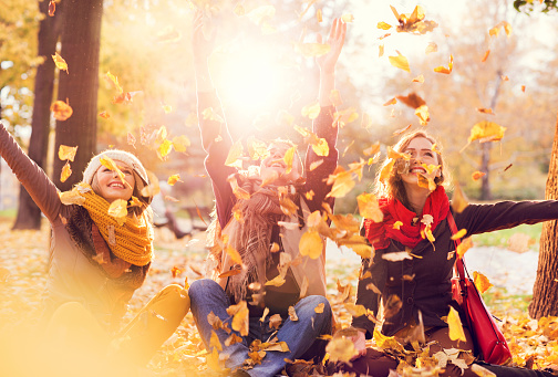 Three teenage friends sitting among autumn leaves in nature and throwing them in the air.