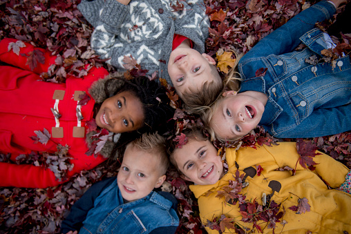 A multi-ethnic group of elementary age children are lying in a circle in a pile of leaves at the park looking up at the camera.